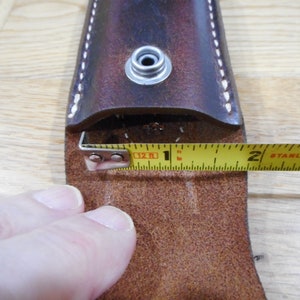 Peut inclure: Un &eacute;tui en cuir marron avec une fermeture &agrave; pression et une boucle en m&eacute;tal. Un m&egrave;tre ruban jaune est utilis&eacute; pour mesurer l'ouverture de l'&eacute;tui, qui mesure 2,5 cm de large.