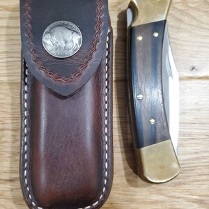Peut inclure: Un &eacute;tui &agrave; couteau en cuir marron avec un bord cousu et un motif de pi&egrave;ce de monnaie en nickel de bison argent&eacute;. &Agrave; c&ocirc;t&eacute; se trouve un couteau pliant avec un manche en laiton et une incrustation en bois fonc&eacute;.