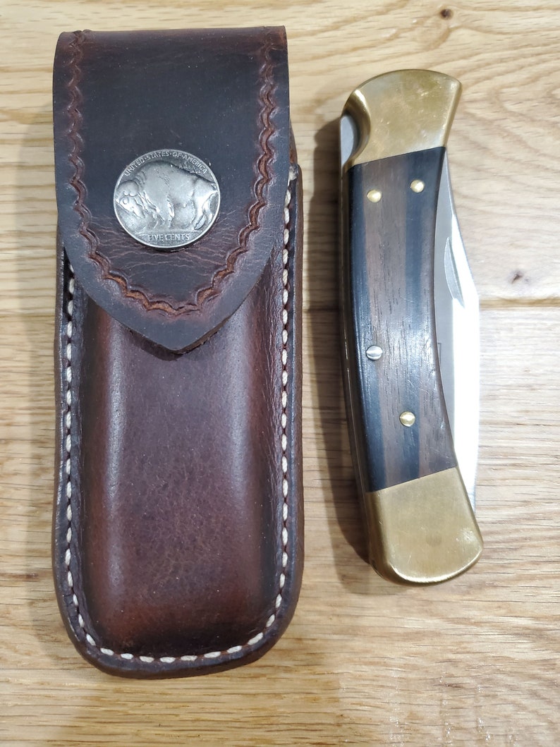 Peut inclure: Un &eacute;tui &agrave; couteau en cuir marron avec un bord cousu et un motif de pi&egrave;ce de monnaie en nickel de bison argent&eacute;. &Agrave; c&ocirc;t&eacute; se trouve un couteau pliant avec un manche en laiton et une incrustation en bois fonc&eacute;.