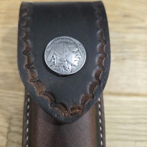 May include: A brown leather knife sheath with a decorative stitching pattern and a silver buffalo nickel coin accent.