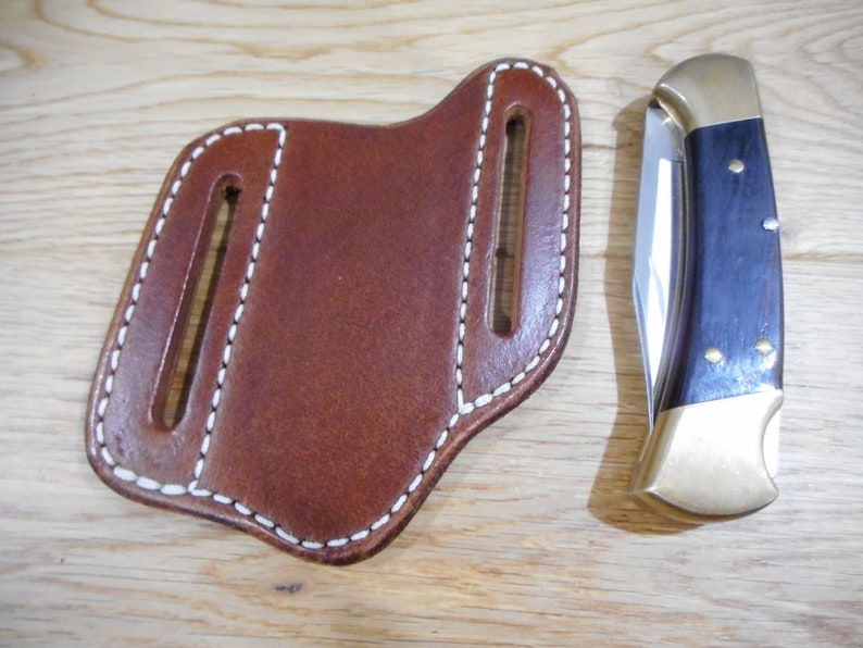 May include: A brown leather knife sheath with white stitching. The sheath has two belt loops and a single slot for a knife. A folding knife with a brown wood handle and a brass bolster is shown next to the sheath.