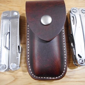 Peut inclure: Deux couteaux multi-outils de poche argentés avec un étui en cuir marron. L'étui est doté d'une couture blanche et d'un bouton argenté.