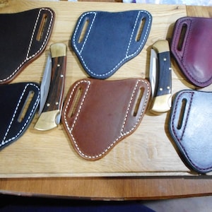 Peut inclure: Six étuis à couteau en cuir de différentes couleurs, dont noir, marron, bleu et bordeaux. Chaque étui possède deux passants de ceinture et des coutures blanches. Deux couteaux de poche avec des manches en bois sont également représentés.