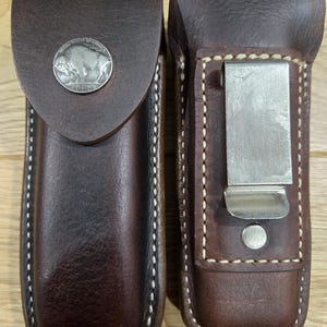 May include: Two brown leather knife sheaths. One sheath features a silver buffalo nickel and a flap closure. The other sheath has a metal clip on the back. Both sheaths have white stitching around the edges.