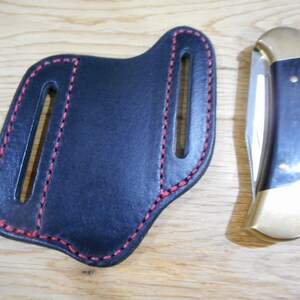 May include: A black leather knife sheath with red stitching. The sheath has two belt loops. A folding knife with a wood handle and a brass bolster is shown next to the sheath.
