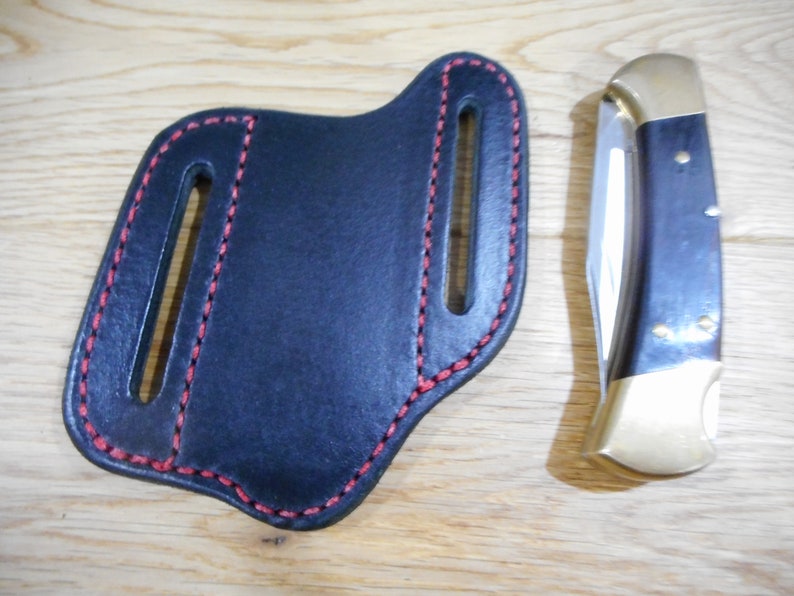 May include: A black leather knife sheath with red stitching. The sheath has two belt loops. A folding knife with a wood handle and a brass bolster is shown next to the sheath.
