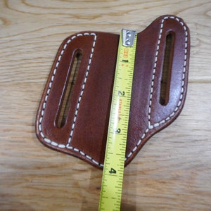 May include: Brown leather knife sheath with white stitching and two slots. The sheath is measured with a tape measure, showing it is approximately 4 inches long.