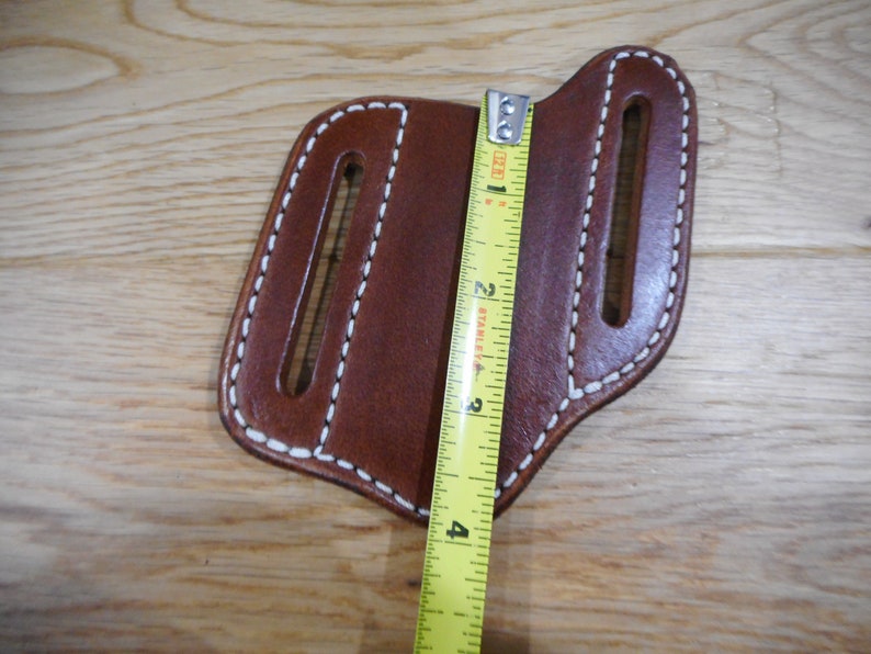 May include: Brown leather knife sheath with white stitching and two slots. The sheath is measured with a tape measure, showing it is approximately 4 inches long.