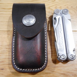 May include: A silver Leatherman multi-tool with a brown leather carrying case. The case has a metal button with a buffalo design.