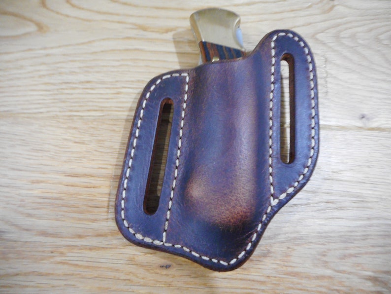 May include: A brown leather knife sheath with white stitching. The sheath has two belt loops and is designed to hold a folding knife.