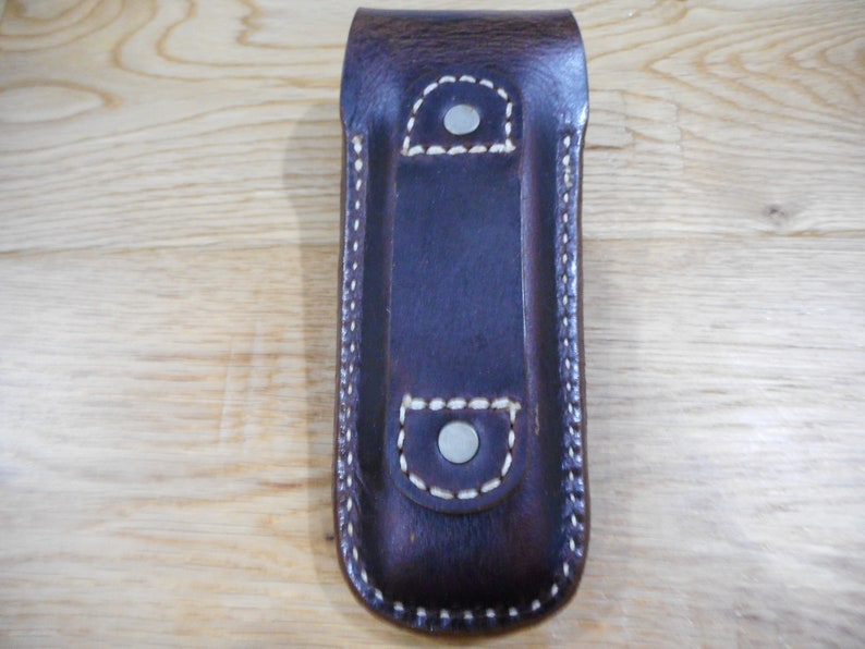 Peut inclure: Un &eacute;tui en cuir marron avec des coutures blanches et des accents m&eacute;talliques argent&eacute;s. L'&eacute;tui a deux poches et est con&ccedil;u pour contenir un couteau de poche.