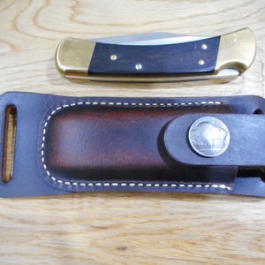 May include: A brown leather knife sheath with a metal buffalo head design. The sheath has a belt loop and a stitched edge.