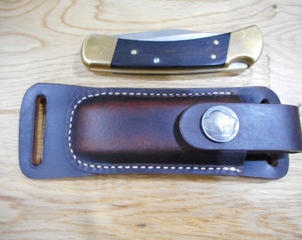 May include: A brown leather knife sheath with a metal buffalo head design. The sheath has a belt loop and a stitched edge.