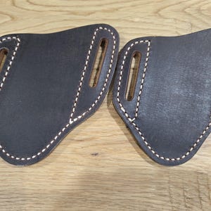 Peut inclure: Deux étuis de ceinture en cuir marron foncé avec une finition lisse et des coutures épaisses de couleur claire sur les bords. Chaque étui possède deux fentes verticales pour la fixation à la ceinture, une forme asymétrique et une structure robuste.