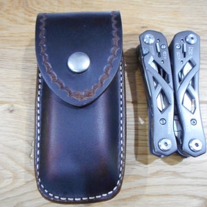 May include: A brown leather case with white stitching and a snap closure. The case is designed to hold a multi-tool. A gray multi-tool with a black handle is shown next to the case.