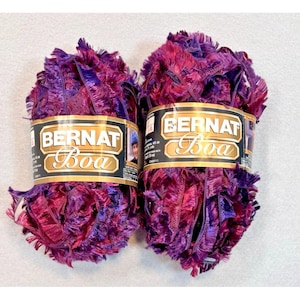May include: Two skeins of Bernat Boa yarn featuring a textured, fringed appearance in variegated shades of deep purple, magenta, and violet. Each skein is wrapped in a gold and black paper label with the text Bernat Boa printed in white.