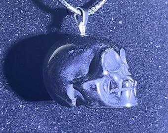 May include: A silver skull pendant necklace with a black obsidian skull charm. The skull has a detailed design with a textured surface.