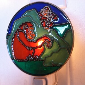 May include: A stained glass nightlight featuring two monkeys, one red and one brown, on a green and blue background.