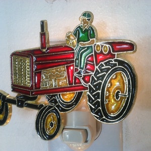 May include: A decorative night light featuring a red tractor with gold and black accents. A figure in green and a helmet is seated on the tractor. The night light has a white base and a switch.