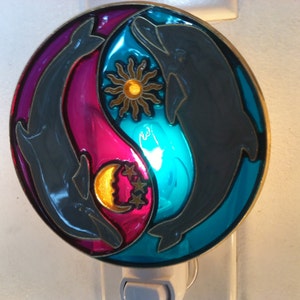 May include: A nightlight featuring a yin yang design with two dolphins swimming in opposite directions. The yin yang symbol is depicted in shades of pink, blue, and turquoise. The dolphins are gray and the background is white.