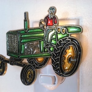 May include: Green and black tractor night light with a gold trim. The tractor has a driver in a red shirt and a white hat. The night light plugs into a standard electrical outlet.