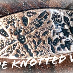 May include: A copper-colored, oval-shaped decorative piece with an embossed floral and insect design. The design includes leaves, flowers, and a dragonfly. The text "THE KNOTTED VINE" is at the bottom. The piece has a metallic finish.