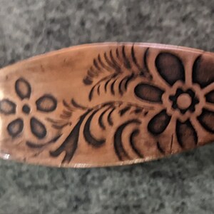 May include: A copper-colored metal bookmark with a floral design. The bookmark is shaped like a long oval and has a dark brown floral pattern.