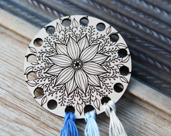 May include: A wooden thread organizer with 13 holes, featuring a black and white floral design. Three strands of embroidery floss are threaded through the holes.