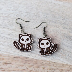 May include: A pair of wooden earrings featuring a cute cartoon cat with a skeleton design. The earrings are brown with white details and have silver hooks.