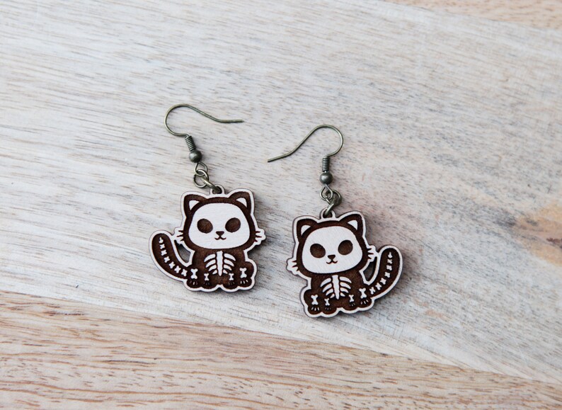 May include: A pair of wooden earrings featuring a cute cartoon cat with a skeleton design. The earrings are brown with white details and have silver hooks.