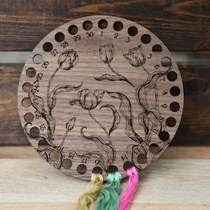 May include: A round wooden embroidery thread organizer with 30 holes numbered 1 to 30. The organizer has a floral design with tulips and leaves. There are four strands of embroidery floss in different colors hanging from the bottom of the organizer.
