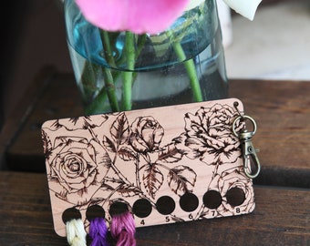 May include: Wooden thread organizer with a floral design featuring roses and leaves. The organizer has eight slots numbered 4 through 8. Three strands of embroidery floss are threaded through the slots.