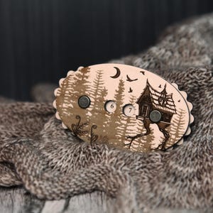 May include: A wooden, oval-shaped magnet with a forest and house design. The magnet has a light brown background with a forest silhouette, a house, a moon, and birds. The numbers 6 and 7 are visible. The magnet is on a gray knitted scarf.