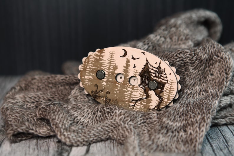 May include: A wooden, oval-shaped magnet with a forest and house design. The magnet has a light brown background with a forest silhouette, a house, a moon, and birds. The numbers 6 and 7 are visible. The magnet is on a gray knitted scarf.