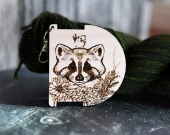 May include: A wooden yarn bowl with a raccoon design. The raccoon is surrounded by flowers and a small bird is perched on its head. The bowl has a metal hook for hanging.