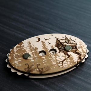 May include: Oval wooden game piece with a forest and house design. The piece has a scalloped edge, two embedded magnets, and the numbers 6 and 7. The artwork includes a moon, birds, and trees.