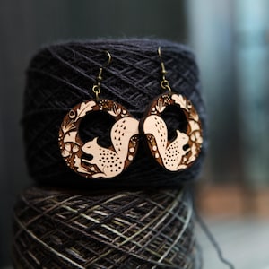 May include: Wooden squirrel earrings with a circular design. The earrings feature a light brown squirrel silhouette surrounded by floral and leaf details. The earrings are hanging in front of a dark gray yarn ball.
