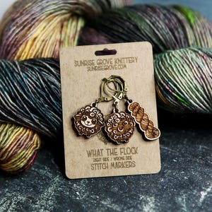May include: Wooden stitch markers with sheep and yarn designs on a kraft paper card. The card reads "WHAT THE FLOCK" and "STITCH MARKERS". Colorful yarn skeins are in the background. The stitch markers are for knitting and crochet projects.