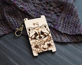 May include: A wooden keychain with a laser-engraved design of a raccoon with flowers. The raccoon is looking at the viewer with a friendly expression. The keychain has a metal ring attached to it.