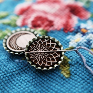 May include: Two round metal bottle caps with intricate designs, one with a crescent moon and the other with a circular pattern, are threaded with a needle and thread on a blue and floral fabric.