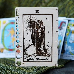 May include: A wooden tarot card with a ruler on the side. The card is a light brown color and features a black and white illustration of The Hermit card from the tarot deck. The card has the Roman numeral IX and the text "The Hermit" printed on it.