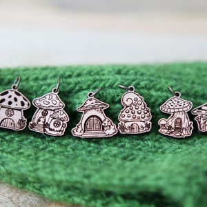 May include: Six wooden mushroom house charms with intricate details, each with a different design. The charms are painted in a light brown color and have a white design. The charms are hanging from a green knitted surface.