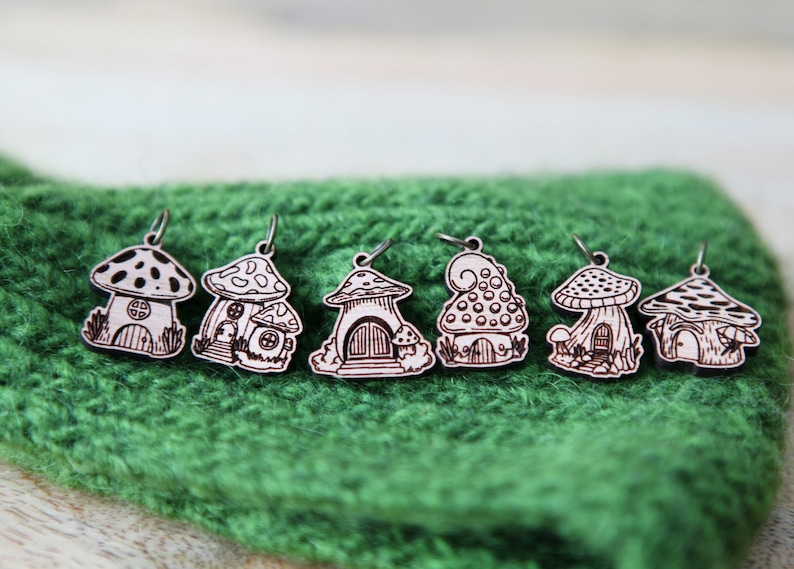 May include: Six wooden mushroom house charms with intricate details, each with a different design. The charms are painted in a light brown color and have a white design. The charms are hanging from a green knitted surface.