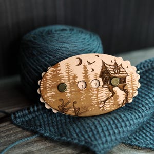 May include: Wooden stitch counter with a forest and house design. The oval-shaped counter has three metal buttons and the numbers 1 and 7. A ball of teal yarn and a knitted piece are in the background.