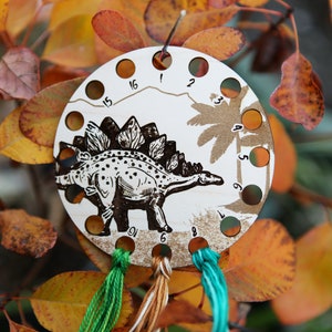 May include: A wooden stitch marker with a dinosaur design and numbered holes. The marker is brown with black details and features a stegosaurus with a spiky back. The numbers on the marker range from 1 to 15.