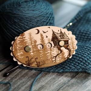 May include: A wooden stitch marker with a forest and house design. The oval-shaped marker has three magnetic closures and is decorated with a crescent moon, birds, and the number "3". The marker is on a blue knitted surface.