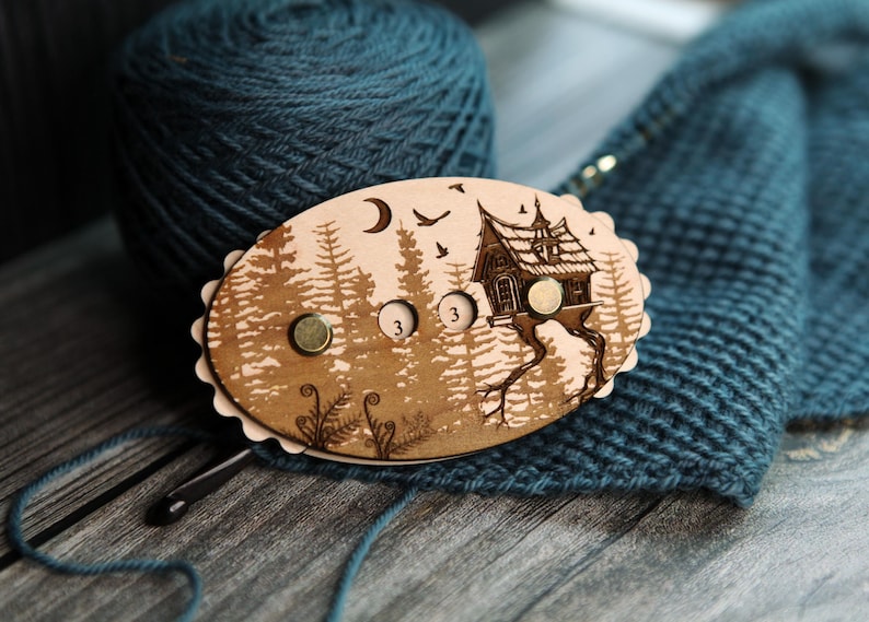 May include: A wooden stitch marker with a forest and house design. The oval-shaped marker has three magnetic closures and is decorated with a crescent moon, birds, and the number "3". The marker is on a blue knitted surface.