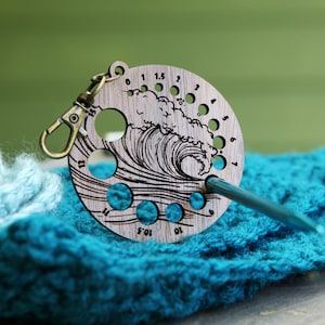 May include: Wooden knitting needle gauge with a wave design and numbered holes. The gauge is attached to a metal ring and is resting on a blue knitted fabric.