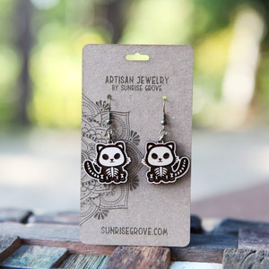 May include: A pair of wooden cat skeleton earrings with silver hooks. The earrings are on a brown card with the text "Artisan Jewelry by Sunrise Grove" and "Sunrisegrove.com".