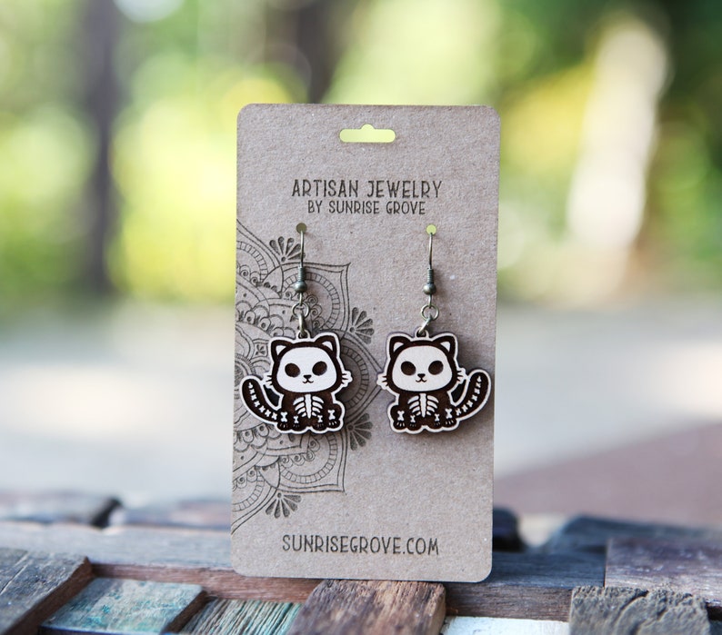 May include: A pair of wooden cat skeleton earrings with silver hooks. The earrings are on a brown card with the text "Artisan Jewelry by Sunrise Grove" and "Sunrisegrove.com".